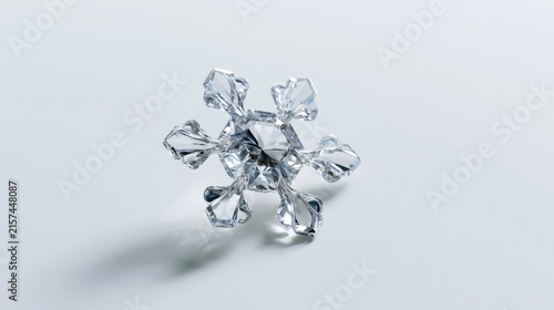 A delicate, crystal snowflake casting a soft shadow on a white surface. The intricate hexagonal structure of the ice crystal is clearly visible