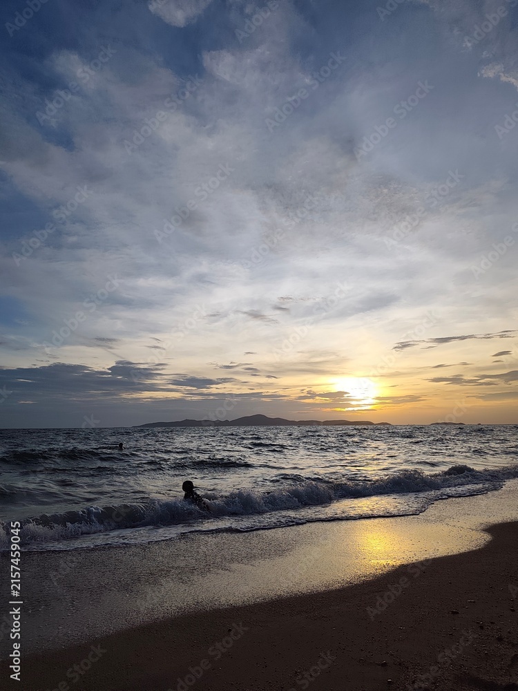 Obraz premium A long vertical shot of a beach sunset with dramatic clouds, a dark sandy shore, small rolling waves, and the silhouette of a lone swimmer in the water near the foreground.
