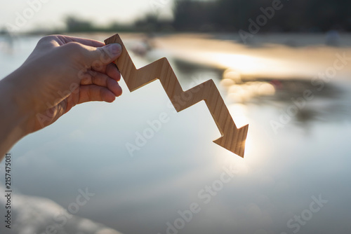 Hand holding wooden downward arrow graph at sunset beach background representing financial crisis, economic loss, market recession, stock crash, business decline, and debt downfall concept.