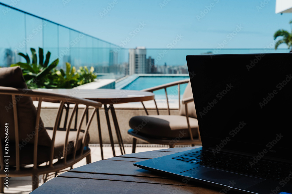Fototapeta premium Laptop on a terrace overlooking a rooftop pool and city skyline. Workation combines remote work with travel for productivity, flexibility and lifestyle balance