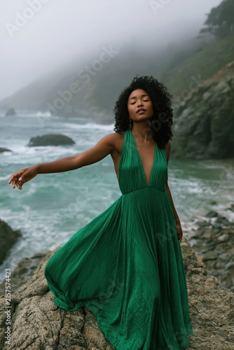 Woman in flowing emerald green dress on a rocky misty coastline for fashion editorial background and dramatic outdoor banner
