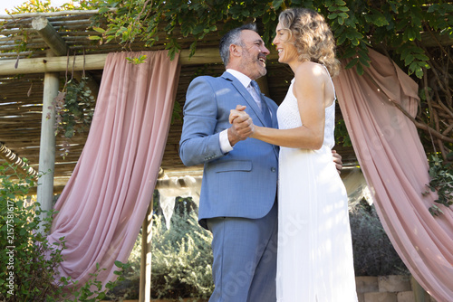 Wedding couple holding hands, dancing in wedding attire under wooden pergola with pink drapes