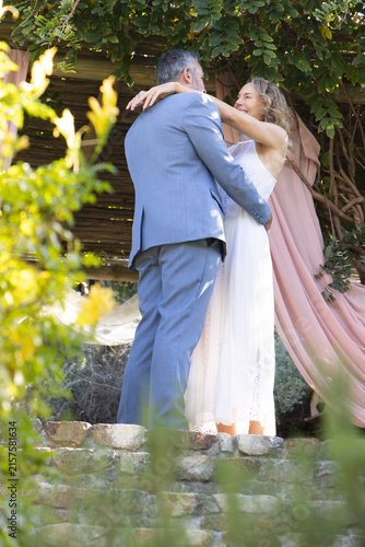 Wedding couple wearing gray suit and lace-dress embracing on low wall under pergola with pink-drape