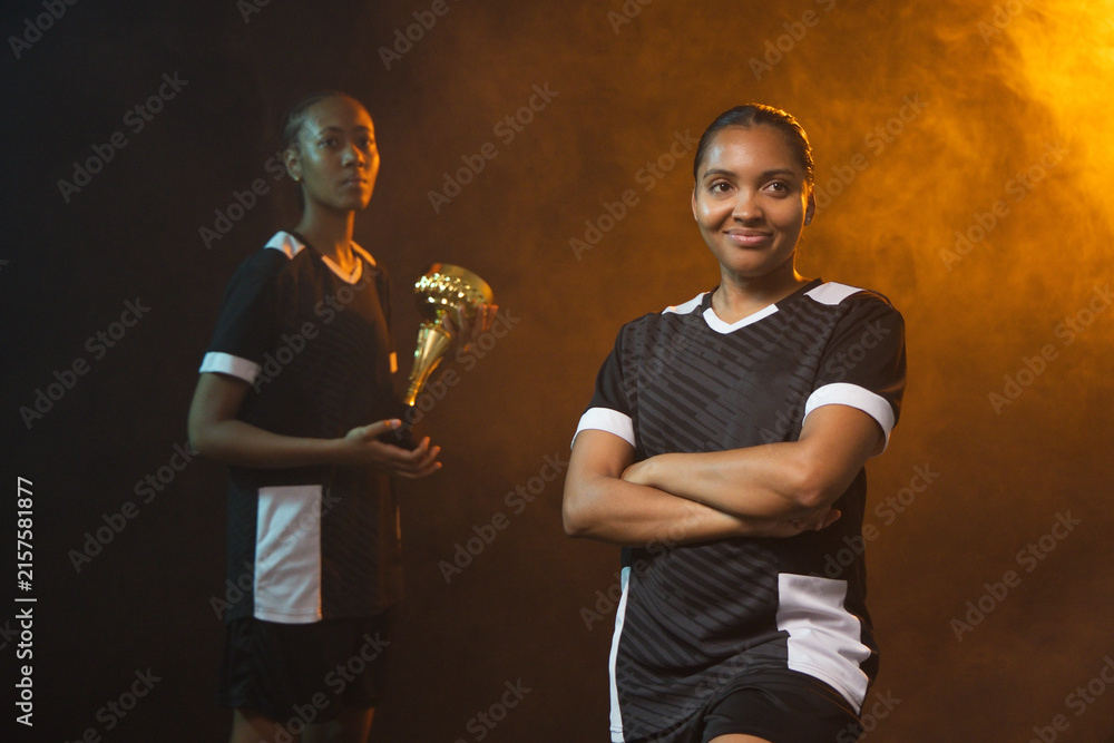 Fototapeta premium African American female team posing in studio, black-white jerseys and shorts, holding gold trophy