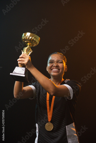 African American woman raising trophy aloft on stage in black jersey, orange-ribbon medal stage-lit
