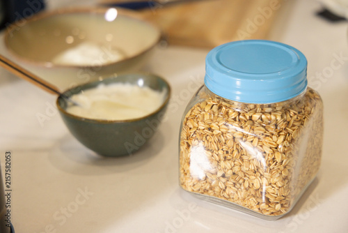 Clear square plastic jar filled with rolled oats sitting on white countertop near bowls, copy space