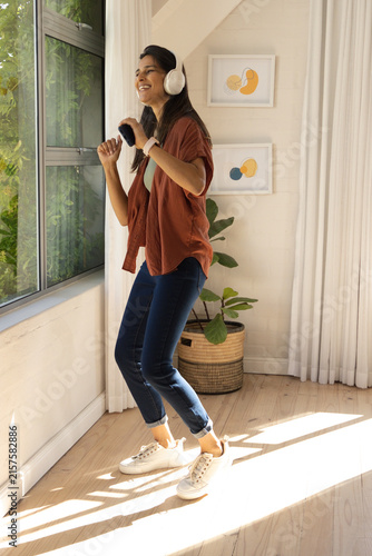 Fotografie Indian woman dancing near large window holding smartphone, wearing white headpho