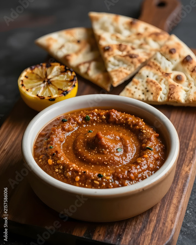 Harissa dip served on a dark wooden slab with a recessed ceramic pool, thick sauce filling the center, charred lemon, roasted garlic and crispy flatbread arranged asymmetrically, extreme close-up