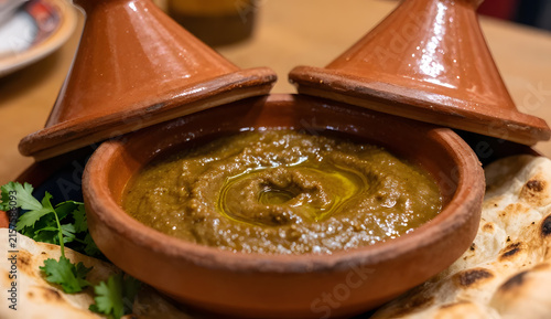 Harissa dip inside a tiny terracotta tagine, lid removed and placed behind the vessel, thick sauce visible inside with olive oil glistening on top, toasted pita and coriander around the base