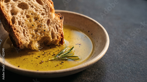 Fresh crusty artisan sourdough bread dipping in extra virgin olive oil with green rosemary herb and black pepper in rustic ceramic bowl on dark table background