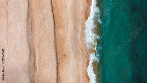 Aerial view of the sandy beach at Nazare with visible tire tracks and turquoise ocean waves crashing on the shore in Nazare, Leiria District, Portugal.