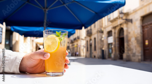 Close up of a hand holding a refreshing lemon cocktail with ice and mint on a sunny outdoor terrace in a European city street, capturing a relaxing summer travel moment.