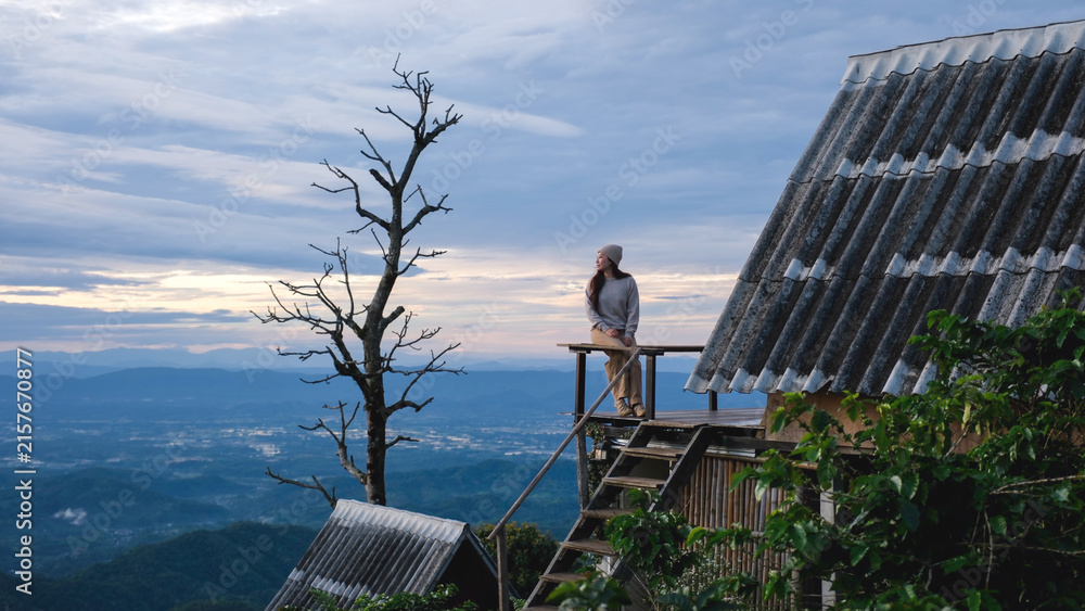 Fototapeta premium A woman sitting on wooden balcony, looking at mountain views on cloudy morning
