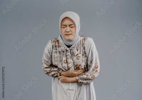 Asian muslim woman in hijab experiencing stomach ache, bending over and holding hands on stomach, discomfort due to menstrual cramps, closing eyes, isolated on white background.