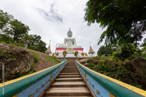 Fotografie Front low angle view of a giant white Buddha statue seated on a lotus base above temple steps under a cloudy sky