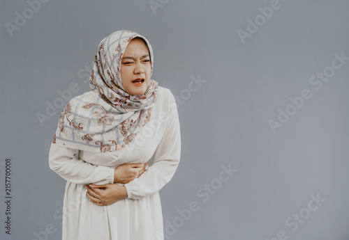 Asian muslim woman in hijab experiencing stomach ache, bending over and holding hands on stomach, discomfort due to menstrual cramps, closing eyes, isolated on white background.