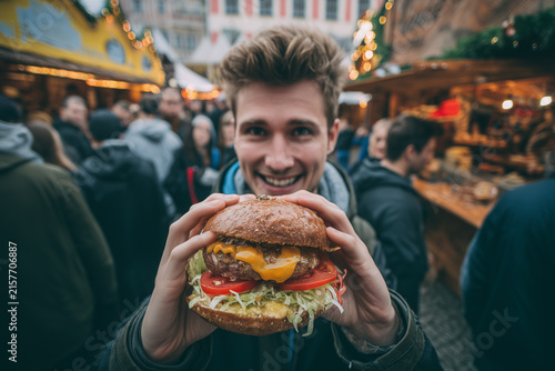 A cheerful young man smiling and holding a giant delicious hamburger with two hands while standing in a lively outdoor evening street food market