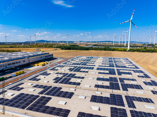 Fotografie Solar panels cover a large flat roof while wind turbines rotate in the backgroun