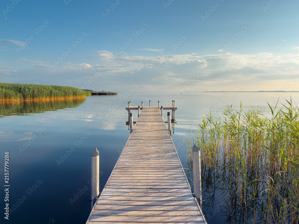 Obraz premium Wooden pier extends into a calm lake, surrounded by reeds and open water under a partly cloudy sky. The tranquil waterfront scene features strong leading lines, reflections, and ample copy space.