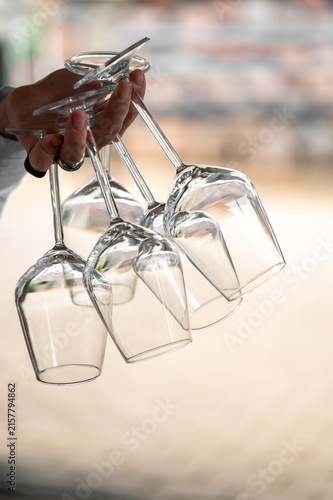 A person holds five upside-down wine glasses by their stems in one hand against a blurred background