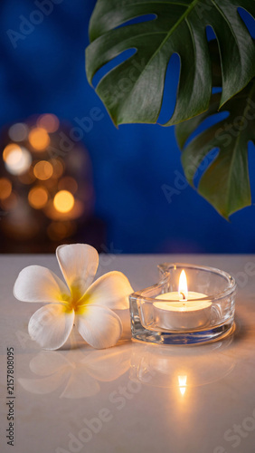 Relaxing spa setting featuring a white tropical plumeria flower and a lit heart shaped glass candle holder on a reflective white surface against a deep blue background with soft bokeh lights.