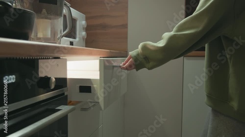 Cropped view of person wearing green sweater reaching to pull out lower drawer beside stainless steel oven, with hand on handle, wooden countertop, white cabinet, and tidy modern kitchen interior