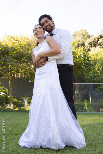 Diverse couple posing on garden lawn, bride wearing white lace wedding gown, groom wearing dark tie