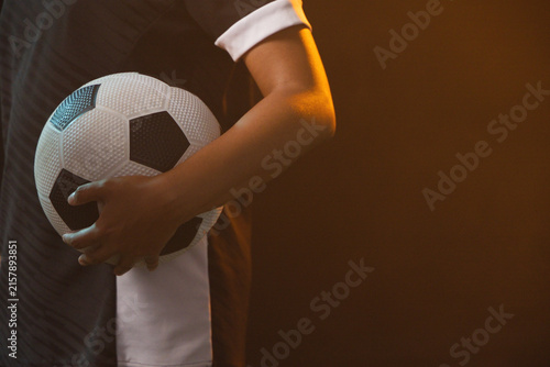 Soccer player is holding black-and-white ball against hip in studio with warm rim light, copy space