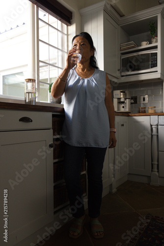 Quadro su tela Senior Asian woman in light blue blouse standing in kitchen drinking from clear
