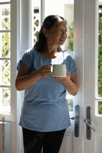 Senior Chinese woman standing by white double doors wearing light-blue shirt and pants, holding mug