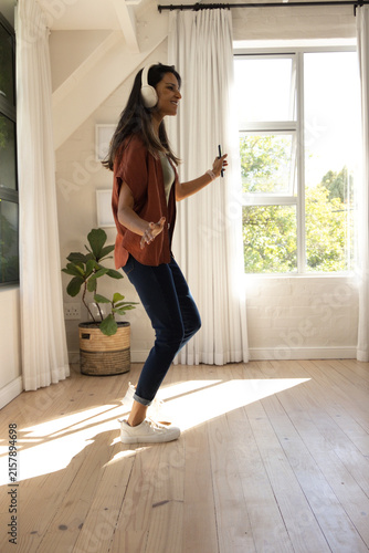 Obraz na płótnie Indian woman dancing in sunlit room at home, wearing headphones and holding smar