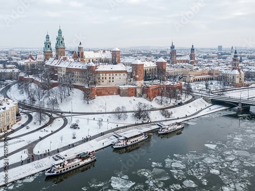 In Krakow, Poland, the Royal Wawel Cathedral and castle lie dusted in winter snow, while the Vistula River hosts tourist boats; an aerial view shows boulevards, promenades, and strolling visitors
