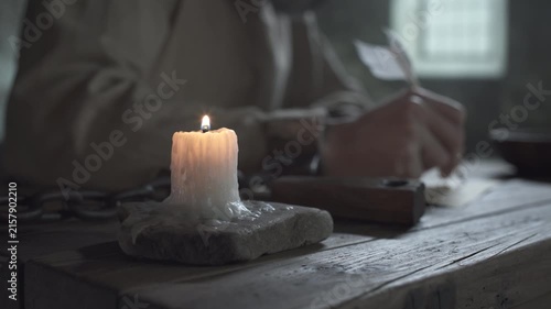 Burning candle on a rustic wooden table against a blurred background of a medieval prisoner in iron shackles writing with a quill pen inside a gloomy cell