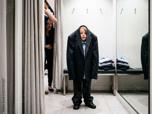 A boy in an oversized school suit is fooling around in a clothing store fitting room with his mother.
