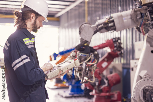 Engineer using pendant controller to program industrial robotic arm in factory. Male technician in hardhat operating automated robot arm in facility. Industrial technician testing mechanical robotic.