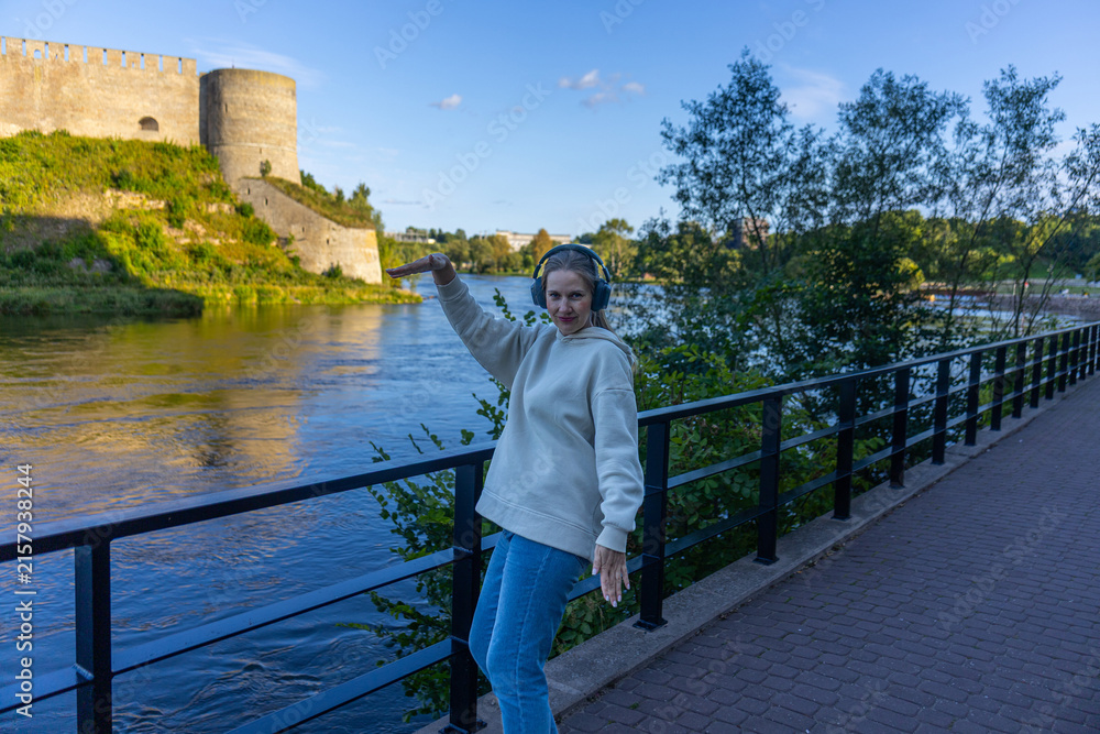 Fototapeta premium Woman with headphones dancing on riverside walkway near fortress