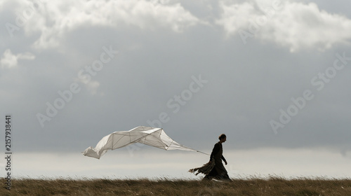 Kite builder carrying a finished geometric cellular kite across an open hillside under a brooding overcast sky, aftermath of a long construction session visible in fabric scraps