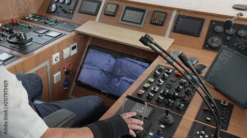 Ship captain operates bridge controls and steering console during vessel maneuvering. CCTV monitor and navigation screens guide boat pilot in harbor. Professional maritime crew workplace.