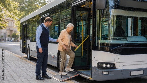 Elderly woman assisted by male bus driver using a wheelchair ramp to board a public transport bus on a city street with trees in the background