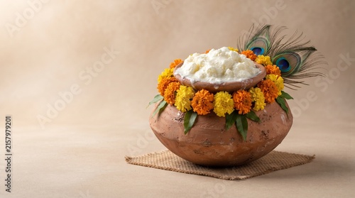 Terracotta matki filled with butter, marigold garland and peacock feather on mat, minimal Janmashtami Krishna offering still life on beige background