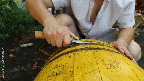 A man scrapes dirt off the surface of an old yellow wooden barrel with a knife. The repairs are taking place on the ground, with removed hoops, tools, and green foliage visible in the background.