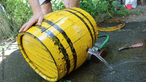 A man holds a yellow wooden barrel, from the side of which water is pouring under pressure. The process takes place in the yard, with a garden hose, tools, and metal hoops lying nearby.
