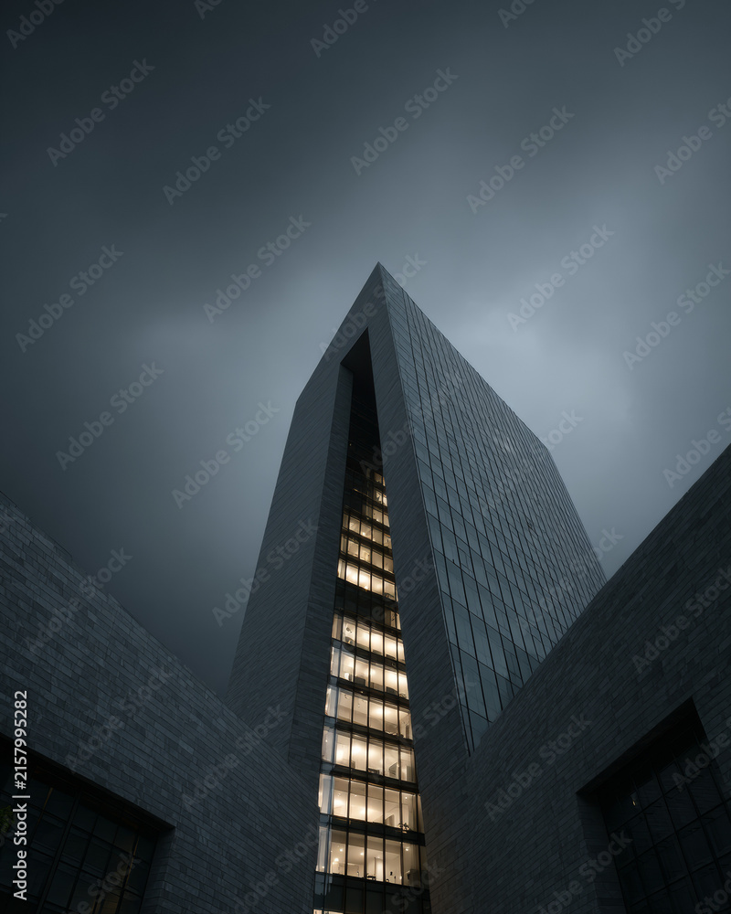 Fototapeta premium Modern glass skyscraper viewed from a low angle under a dark overcast storm sky