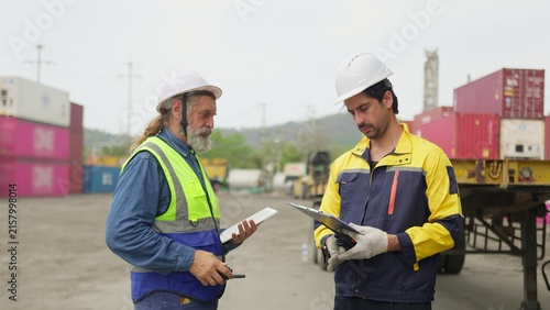 Quadro su tela Two logistics workers discuss a shipping manifest on a clipboard in a container yard, collaborating on supply chain management and cargo inspection, reflecting teamwork and modern workplace themes