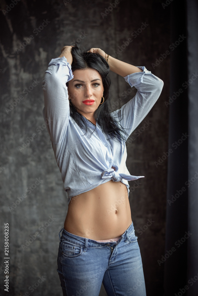 Fototapeta premium An attractive woman is standing in the studio. She is wearing a cropped shirt tied at the waist. Blue jeans fit her figure perfectly, drawing attention to her thin waist.