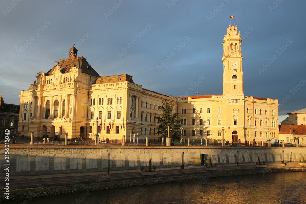 Obraz premium Historic City Hall Palace of Oradea, Romania, illuminated by warm evening sunlight beside the Crișul Repede River under a dramatic dark sky.