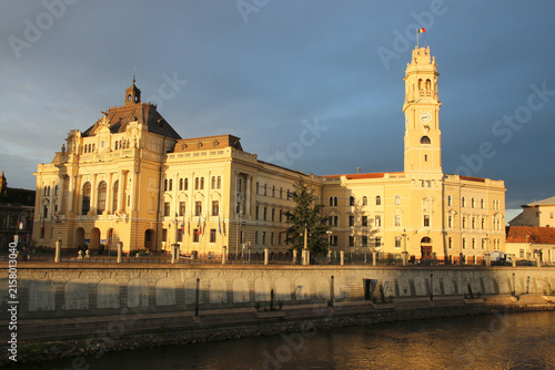 Photography Historic City Hall Palace of Oradea, Romania, illuminated by warm evening sunlight beside the Crișul Repede River under a dramatic dark sky