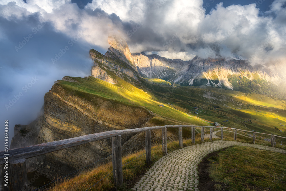 Naklejka premium Seceda mountain peak in clouds with hiking trail and wooden fence in Dolomites, Italy. Summer alpine landscape with sunbeams, green grass meadow slope, high cliffs, dramatic sky and fog at sunset