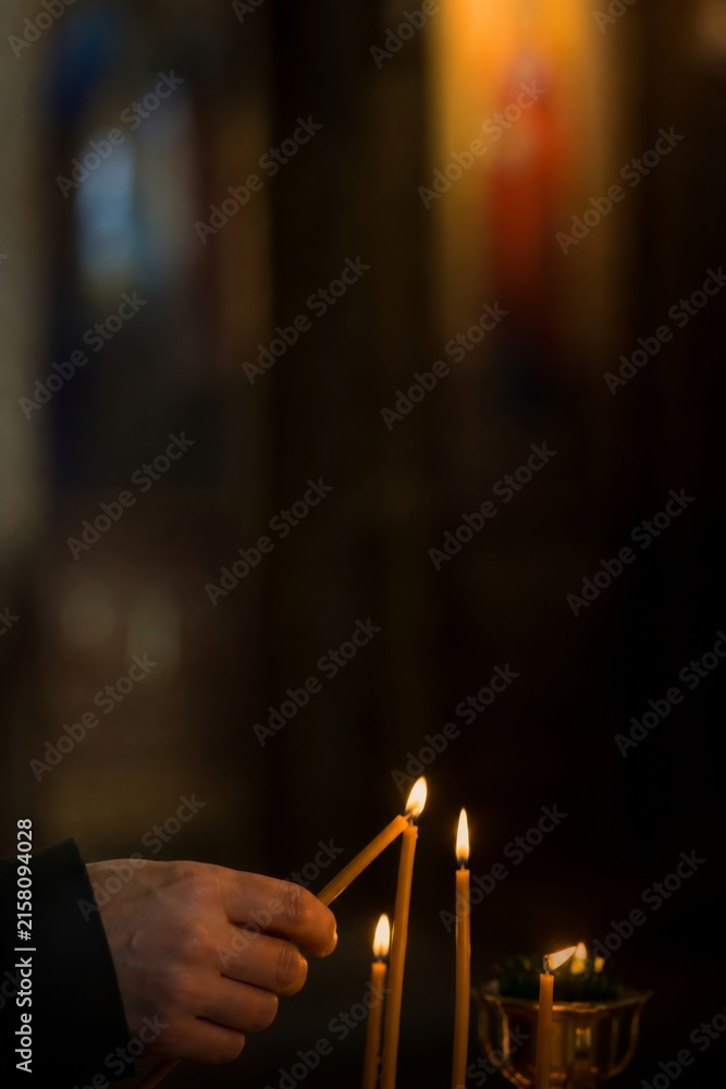 Naklejka premium Hand lighting a candle beside burning candles in a dark interior. Spiritual ritual, remembrance, prayer, and solemn memorial concept with atmospheric copy space.