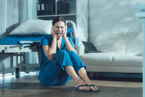 Canvas Print Overwhelmed Nurse in Crisis: A young nurse in blue scrubs sits on the floor of a hospital room, overwhelmed by emotion and covering her ears